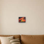 Where the sun becomes a beast canvas print by Artis Berkmanis, the black silhouette of a stag calling against a fiery red and yellow sunset sky. Shown at 25 x 20 cm above a sofa in a Scandinavian living room.