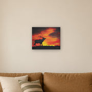 Where the sun becomes a beast canvas print by Artis Berkmanis, the black silhouette of a stag calling against a fiery red and yellow sunset sky. Shown at 51 x 41 cm above a sofa in a Scandinavian living room.