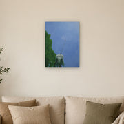 I am a beginning, not an ending canvas print by Artis Berkmanis, a single dandelion seed lifting off into a clear blue sky. Shown at 46 x 61 cm above a sofa in a Scandinavian living room.
