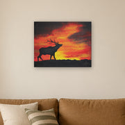 Where the sun becomes a beast canvas print by Artis Berkmanis, the black silhouette of a stag calling against a fiery red and yellow sunset sky. Shown at 76 x 61 cm above a sofa in a Scandinavian living room.
