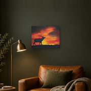 Where the sun becomes a beast canvas print by Artis Berkmanis, the black silhouette of a stag calling against a fiery red and yellow sunset sky. Shown at 51 x 41 cm on a dark wall in a cosy living room.