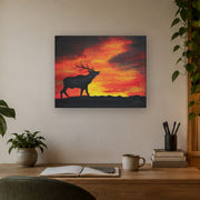 Where the sun becomes a beast canvas print by Artis Berkmanis, the black silhouette of a stag calling against a fiery red and yellow sunset sky. Shown at 51 x 41 cm above a desk in a Scandinavian home office.